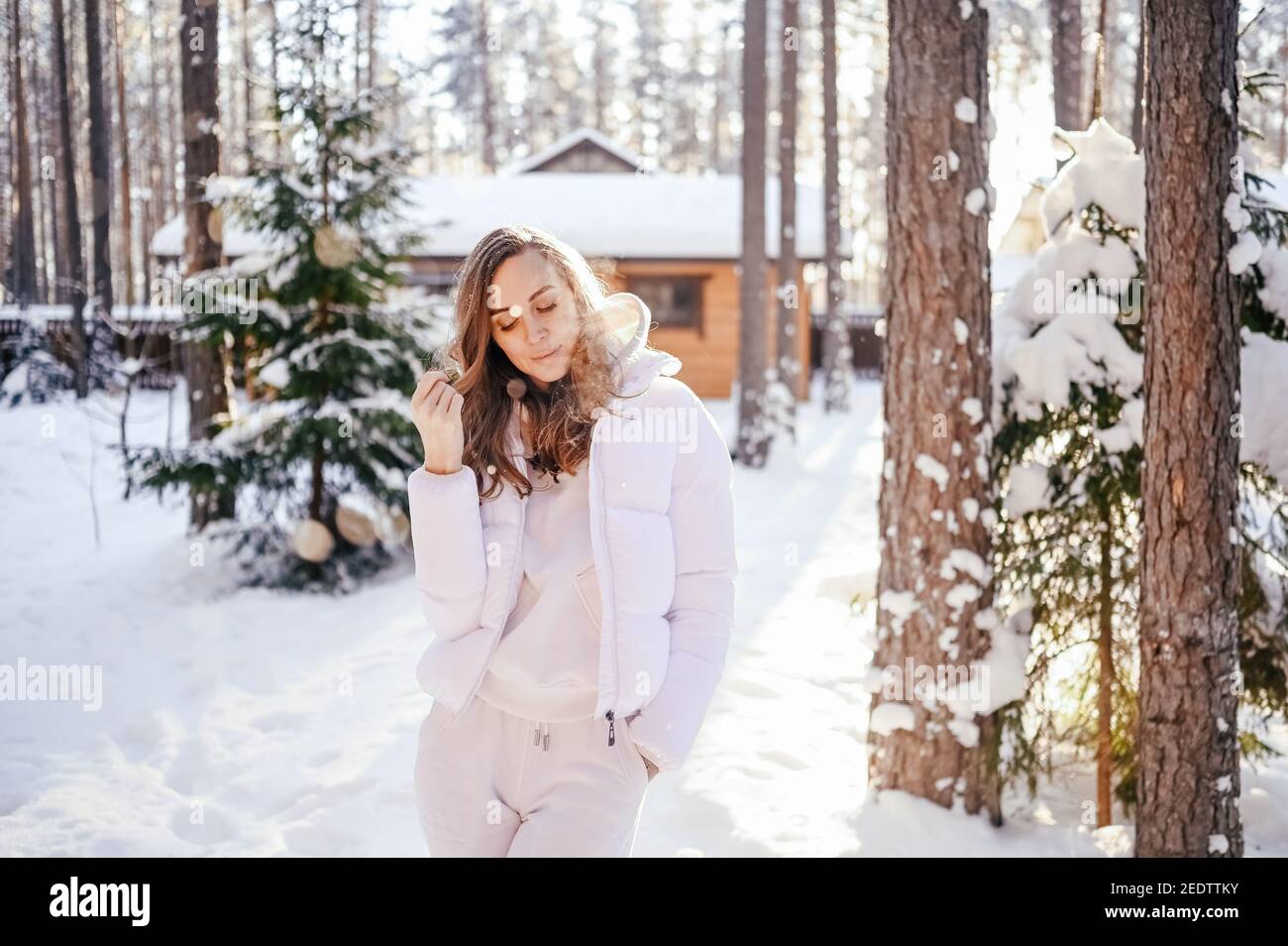 Winter sonnig kalt Porträt im Schneefall der schönen jungen Frau In einer weißen warmen outwear Daunenjacke im Hinterhof Landhaus Holzhaus auf dem Hintergrund der grünen Fichtenkiefer Bäume mit Schnee bedeckt Stockfoto
