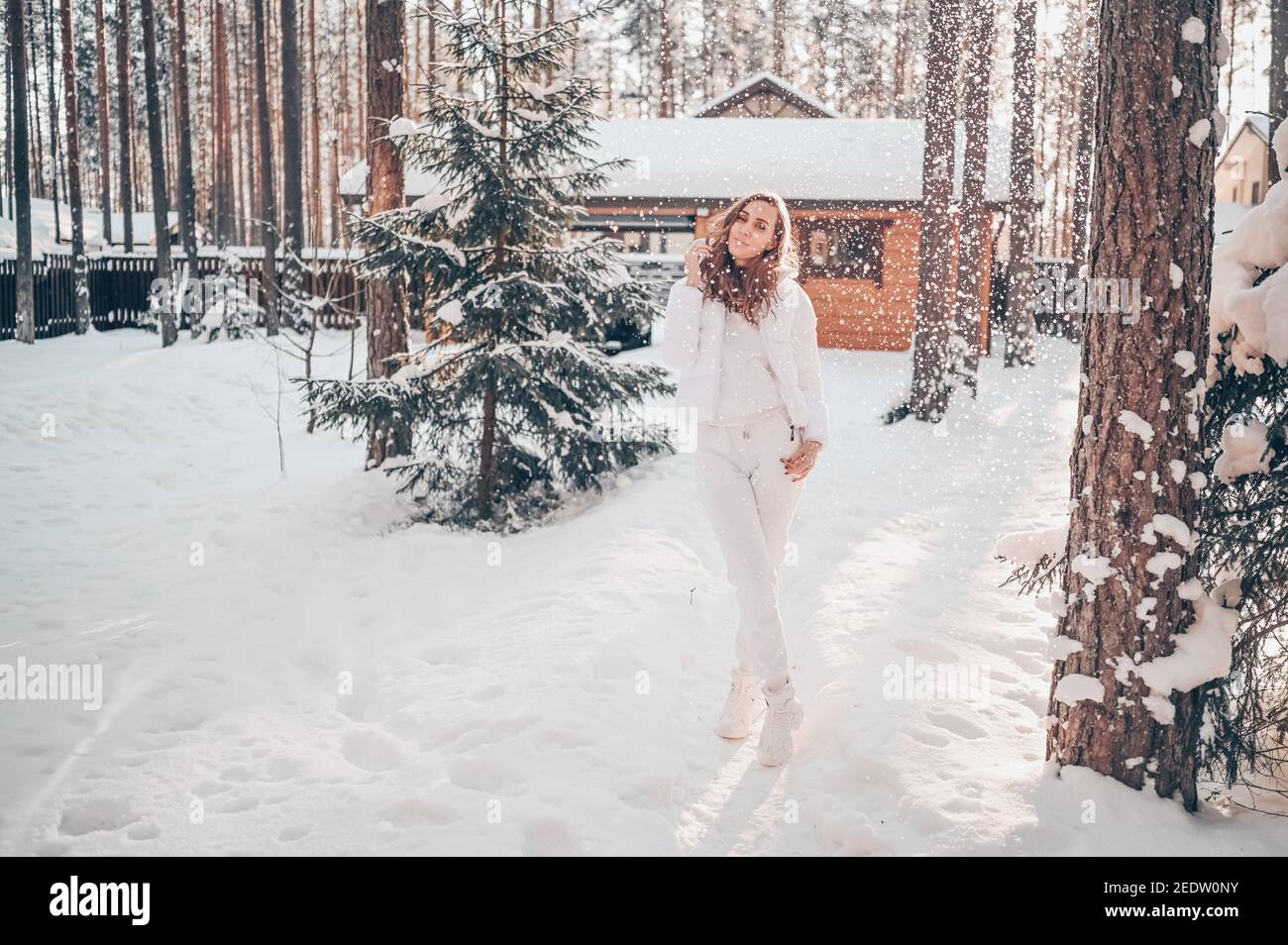 Winter sonnig kalt Porträt im Schneefall der schönen jungen Frau In einer weißen warmen outwear Daunenjacke im Hinterhof Landhaus Holzhaus auf dem Hintergrund der grünen Fichtenkiefer Bäume mit Schnee bedeckt Stockfoto