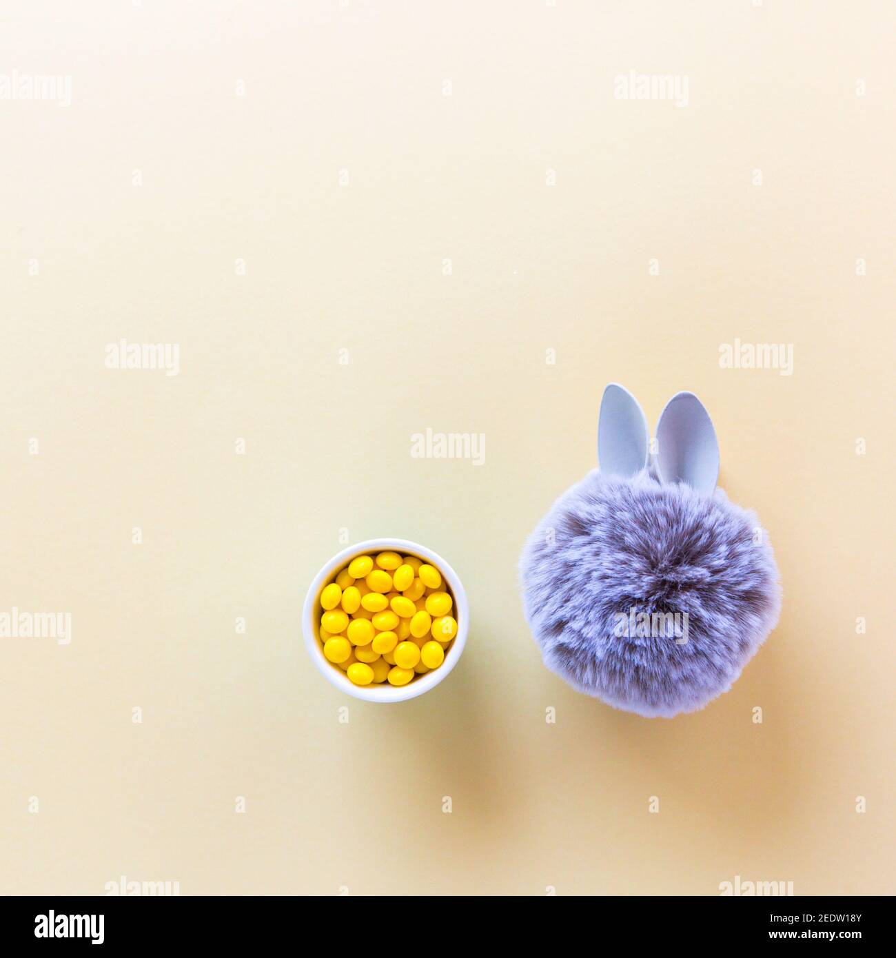 Pelzkugel mit Hase Kaninchenohren mit gelben Bonbons auf hellem Hintergrund. Ostern Minimal Konzept. Flach liegend Stockfoto