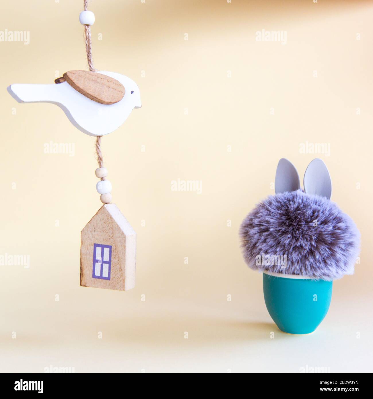 Pelzkugel mit Hasenohren im Eierständer und Holzhaus und Vogel auf pastellfarbenem Hintergrund. Ostern Minimal Konzept. Nahaufnahme Stockfoto
