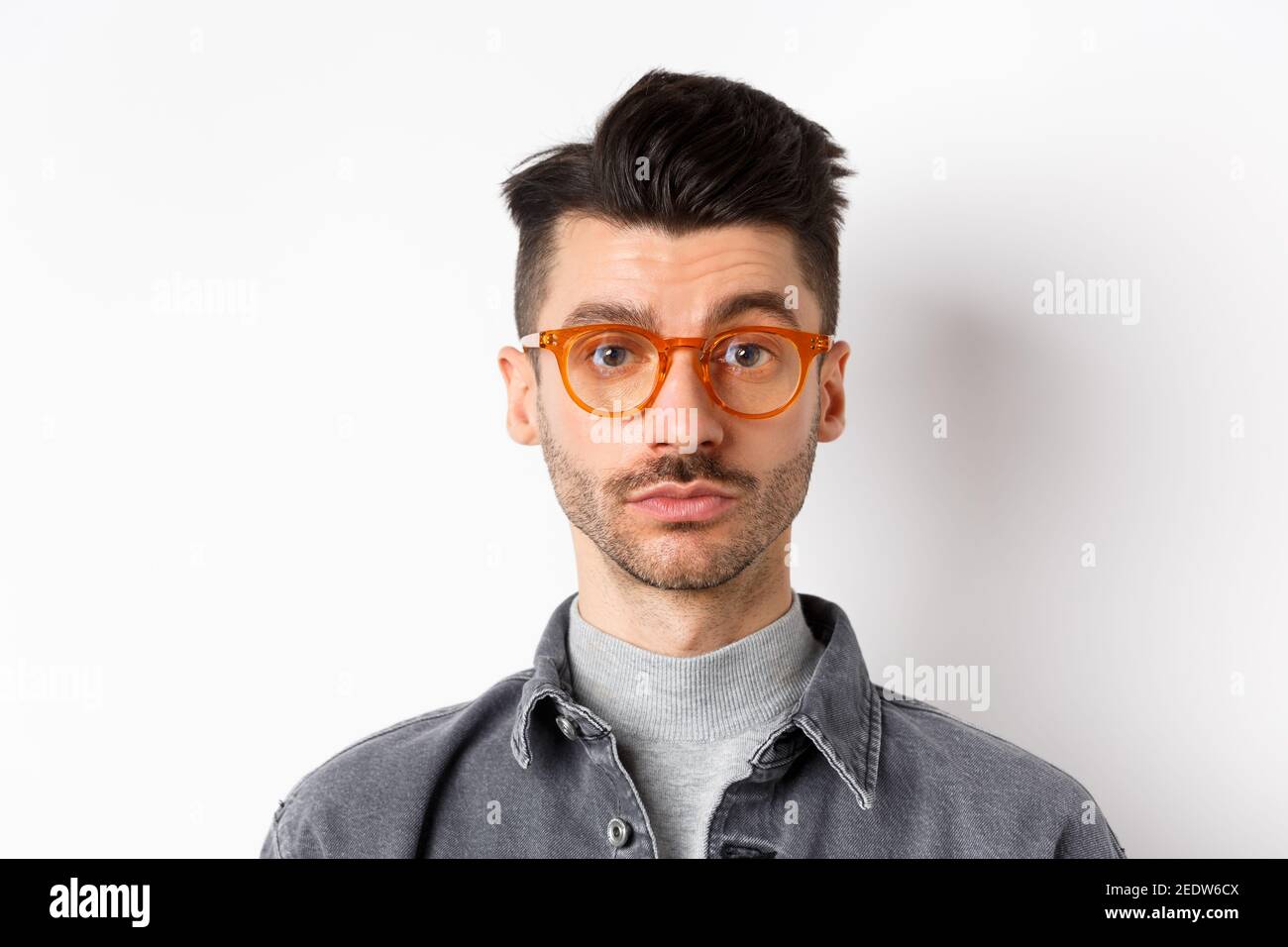 Attraktive Brünette Mann mit Schnurrbart, trägt stilvolle Brille und sehen ernst an der Kamera, keine Emotion pokerface, vor weißem Hintergrund stehen Stockfoto
