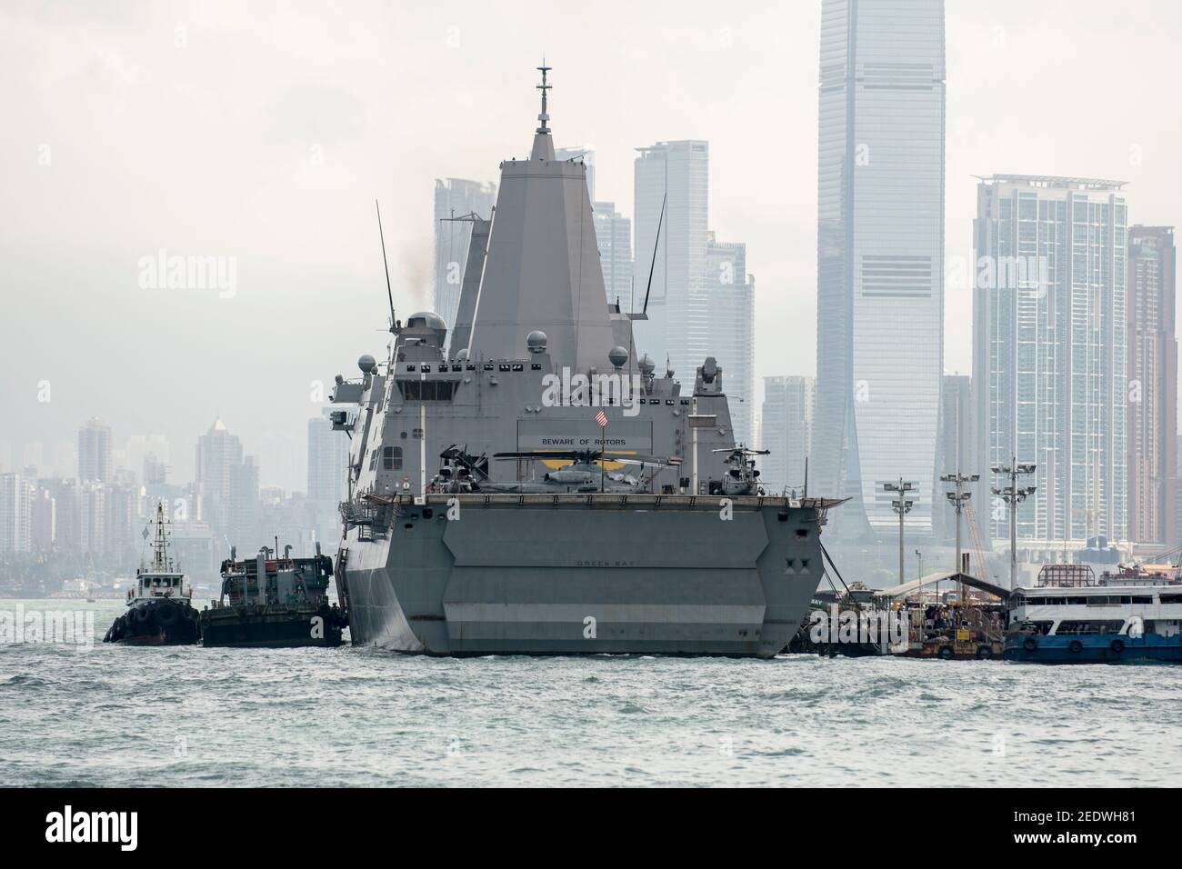 Blick auf das Amphibiendock-Landungsschiff USS Green Bay (LPD 20) vor der Skyline von Gebäuden, angedockt am Eingang zum Victoria Harbour im Rahmen eines planmäßigen Hafenbesuchs. Green Bay ist Teil der Bonhomme Richard Expeditionary Strike Group, die hier in Hongkong, Hongkong, SAR, China, VR China, zu sehen ist. © Time-Snaps Stockfoto