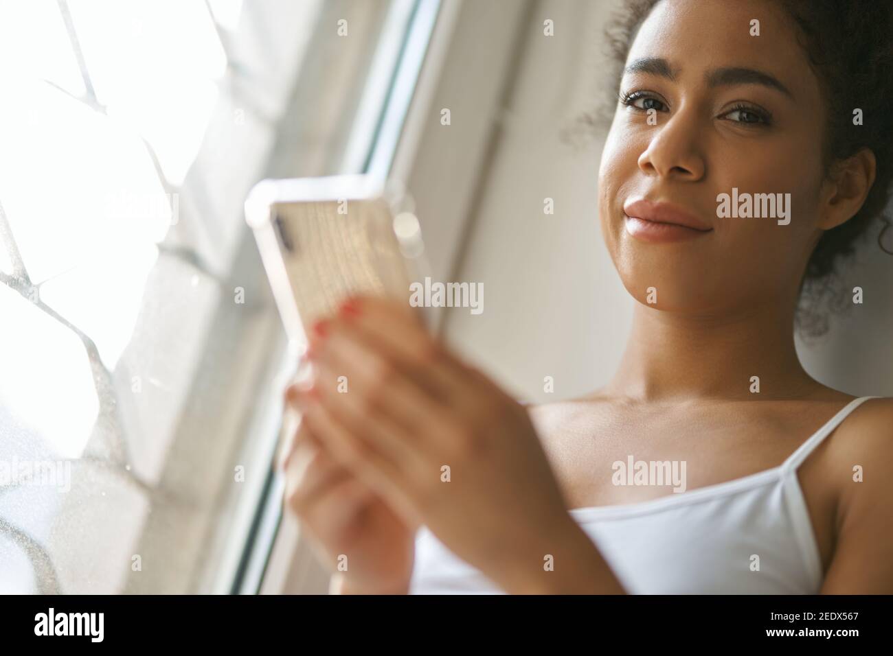 Porträt einer jungen Frau mit gemischter Rasse, die Kamera anschaut, Smartphone benutzt und sich gemütlich fühlt, während sie zu Hause am Fenster sitzt. Lifestyle, Freizeitkonzept Stockfoto Porträt einer jungen Frau mit gemischter Rasse, die Kamera anschaut, Smartphone benutzt und sich gemütlich fühlt, während sie zu Hause am Fenster sitzt. Lifestyle, Freizeitkonzept Stockfoto