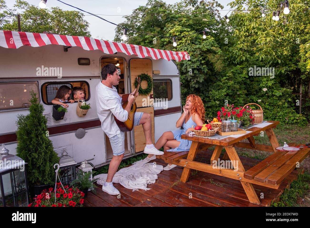 Junger Vater spielt Mädchen Gitarre. Rothaarige Frau Mutter hört sich das Lied an, lacht. Tochter und Sohn schauen aus dem Fenster des Anhängers. Sommer-Spaß t Stockfoto