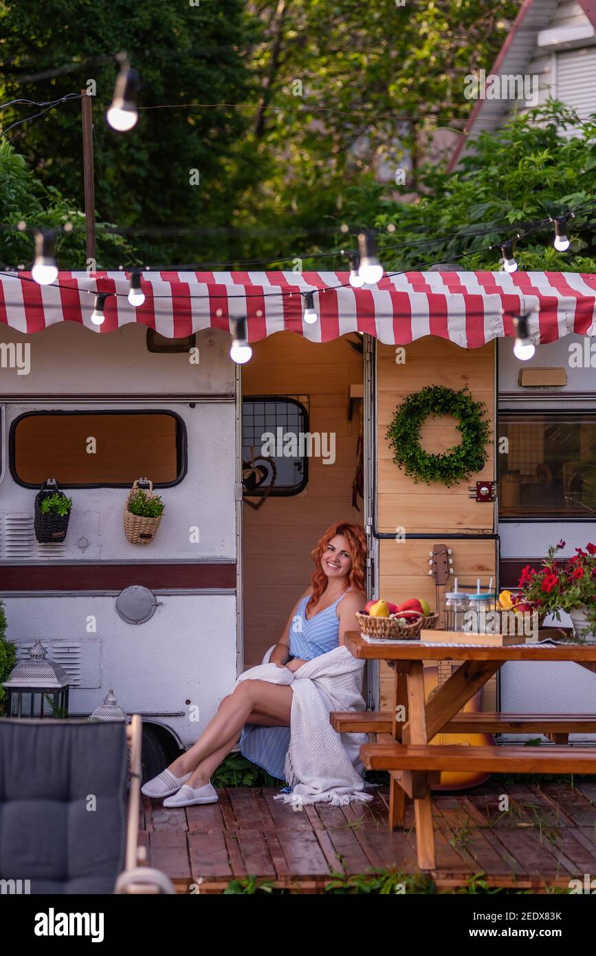 Ein schönes schlankes Mädchen mit roten Lockenhaaren und einem wunderschönen Lächeln in einer blauen Sundress sitzt in der Tür ihres Anhängers. Eine zärtliche Mutter reist mit Stockfoto