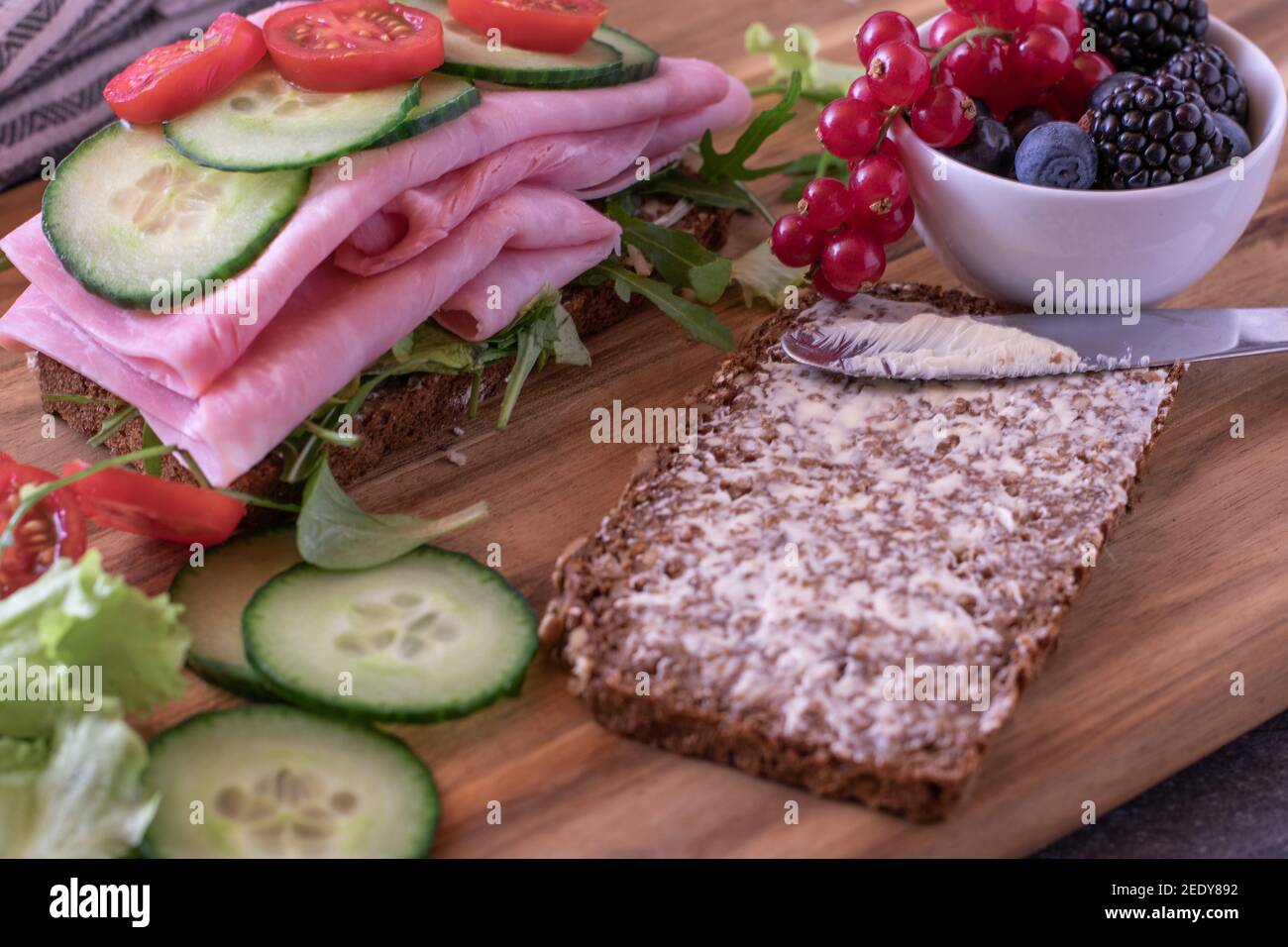 Offenes Sandwich mit Schinken, Tomaten und Gurken, serviert mit frischen Beeren auf einem Holzbrett Stockfoto