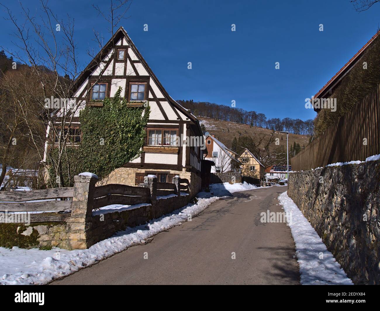 Historisches Fachwerkhaus mit Holzzaun, Steinmauer und asphaltierter Straße im kleinen ländlichen Dorf Gundelfingen, Teil von Münsingen, Deutschland. Stockfoto
