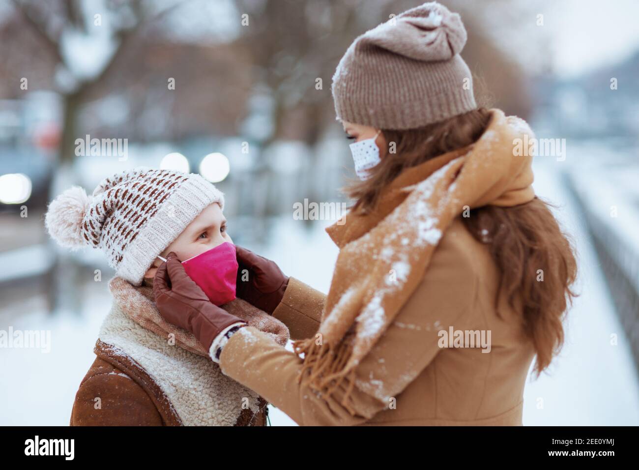 Elegante Mutter und Kind in einem gestrickten Hüte und Mäntel draußen in der Stadt im Winter tragen medizinische Maske. Stockfoto