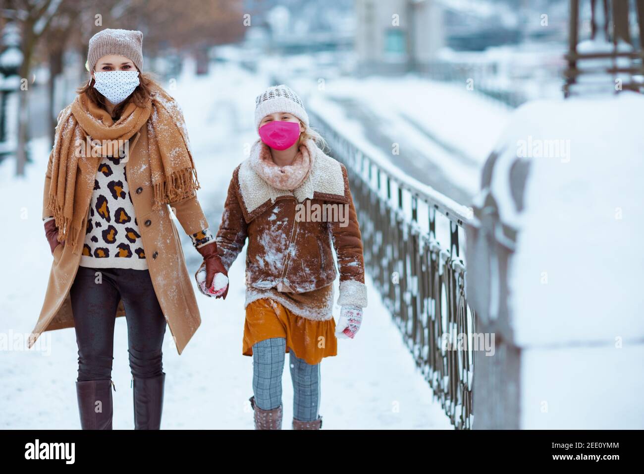 Moderne Mutter und Tochter in einem gestrickten Hüte und Mäntel mit medizinischer Maske zu Fuß außerhalb in der Stadt im Winter. Stockfoto