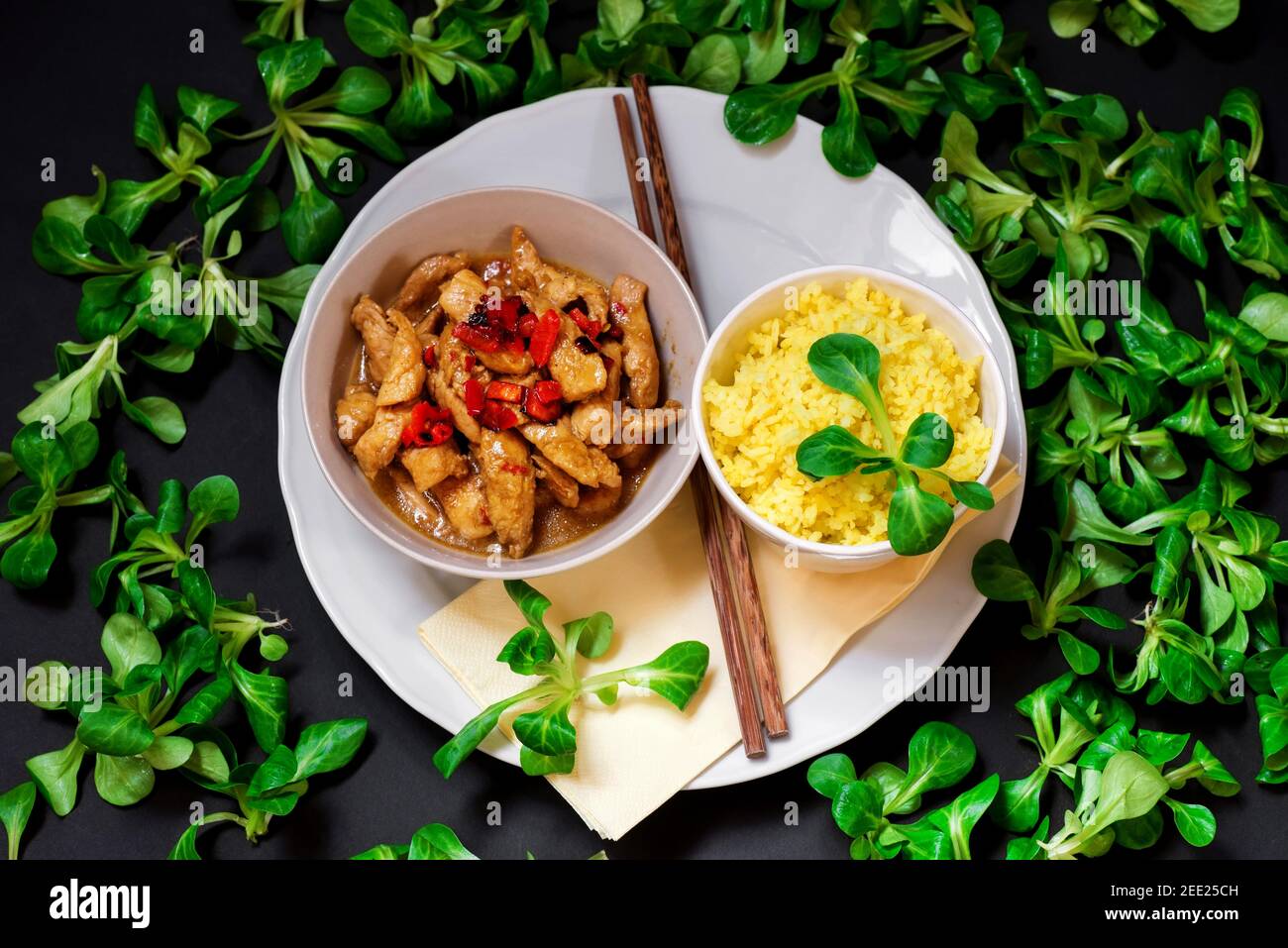 Putenfleischstreifen mit rotem Pfeffer, Curryreis auf weißem Teller, Essstäbchen und vielen Lammsalatblättern ´s schwarzem Hintergrund. Asiatische Küche. Stockfoto