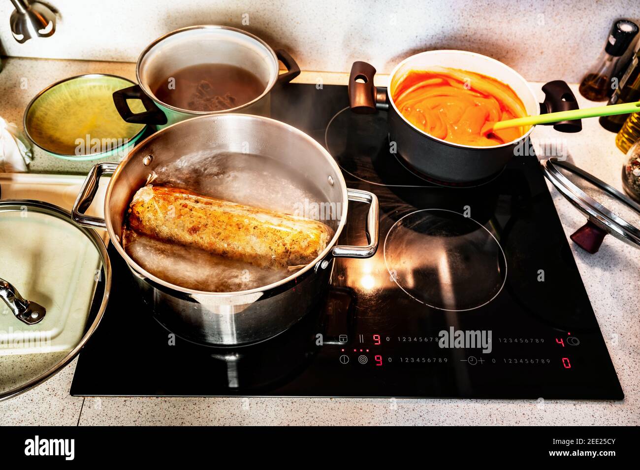Preparatio Essen auf heißer Küche Keramik-Kochfeld. Knödel in kochendem Wasser im großen Topf, Deckel, Tomatensauce in kleinem Topf mit Löffel, Rindfleisch in Brühe. Stockfoto