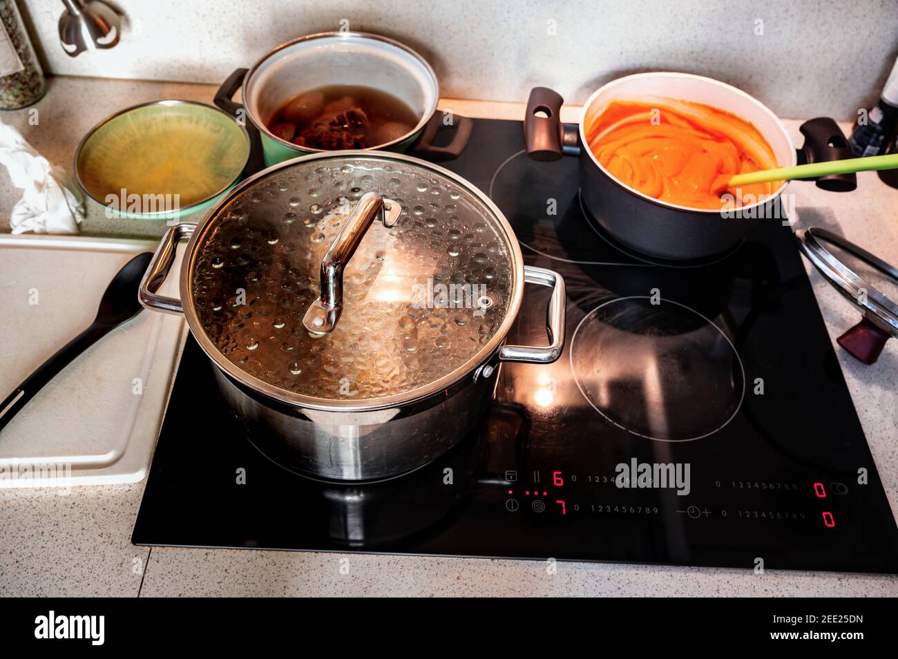 Zubereitung von Mahlzeiten auf heißer Küche Keramik-Kochfeld. Kochendes Wasser im großen Topf mit Deckel, Tomatensauce im Topf, Löffel, Fleisch in Brühe im Topf, Küchenbrett, Schlepptau Stockfoto