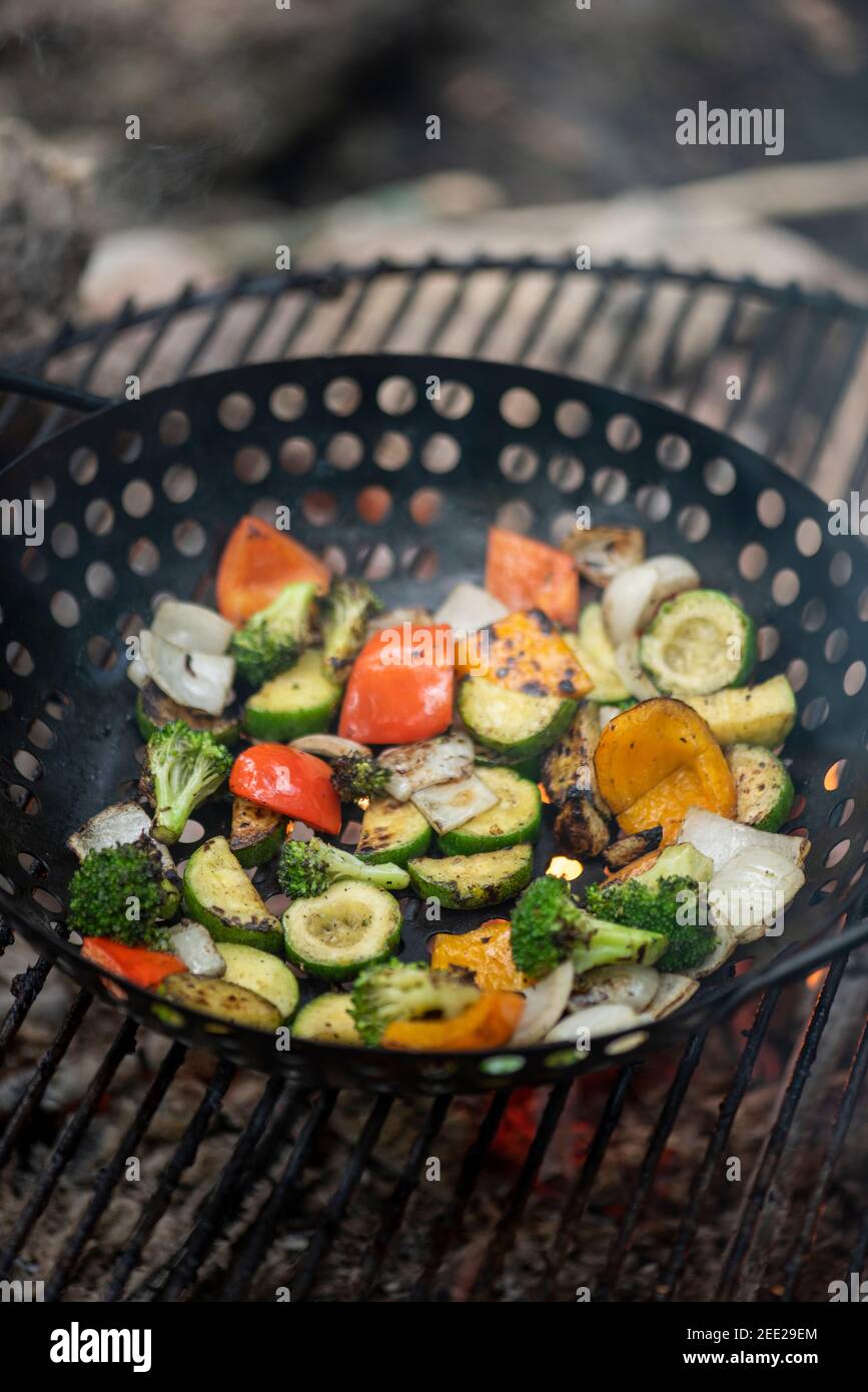 Ein Korb mit Gemüse und marinierten Hähnchenschenkeln kochen auf einem heißen Lagerfeuer. Essen im Freien. Gegrilltes Gemüse. Essen auf einem Campingausflug Stockfoto