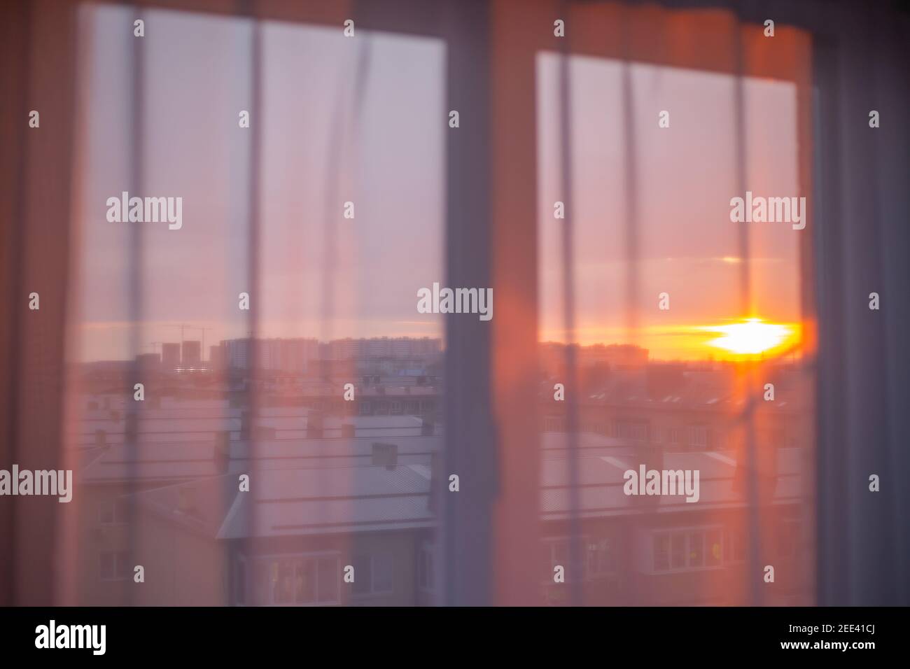 Die Strahlen der Morgensonne fallen durch den weißen Tüll auf dem Fenster. Sonnenaufgang über der Stadt im Hintergrund. Neuer Tag, stimmungsvolle Stimmung. Stockfoto