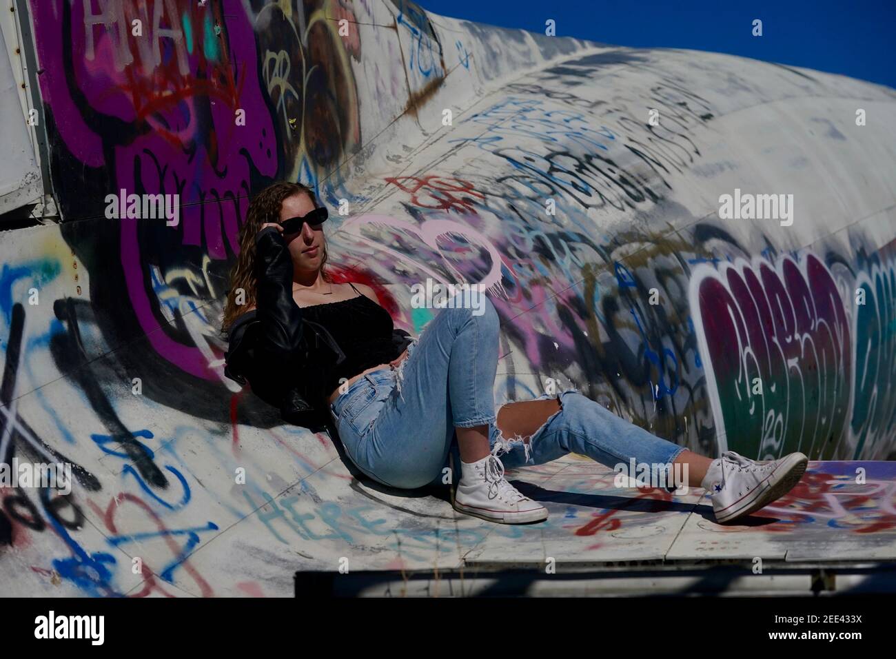 Ein großes blondes Teenager-Mädchen trägt zerrissene Jeans und ein Weiße Tank Top Posen für Porträts von verlassenen Flugzeugen bedeckt In Graffiti in der Wüste wild Stockfoto
