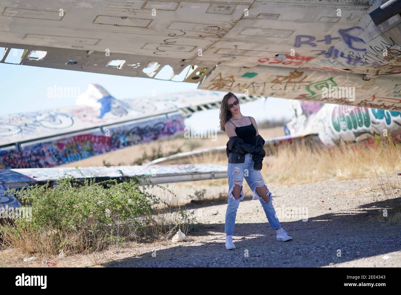 Ein großes blondes Teenager-Mädchen trägt zerrissene Jeans und ein Weiße Tank Top Posen für Porträts von verlassenen Flugzeugen bedeckt In Graffiti in der Wüste wild Stockfoto