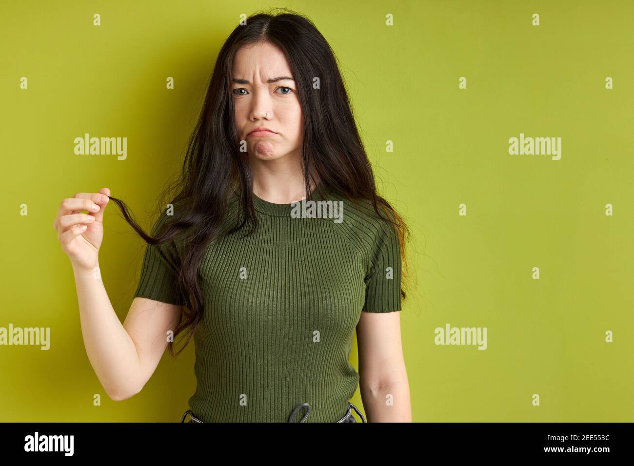 Nachdenkliche Brünette Frau schaut atcamera, während sie Haarsträhne in den Händen hält, müssen ausschneiden, traurige Emotionen fühlen, isolierter grüner Hintergrund Stockfoto