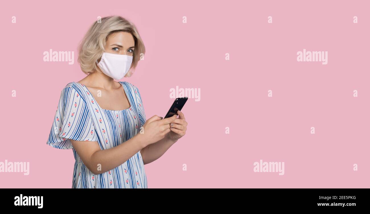 Frau mit medizinischer Maske im Gesicht und blonden Haaren ist Chatten auf Handy auf einer rosa Studio Wand mit kostenlos Platz Stockfoto