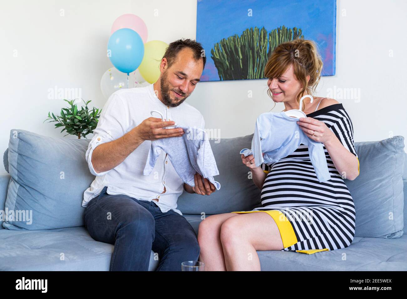 Ein junges Ehepaar sitzt auf der Couch im Wohnzimmer. Frau ist schwanger und lächelt, wie sie auf die Kleidung schauen, für das Baby sind sie ex Stockfoto