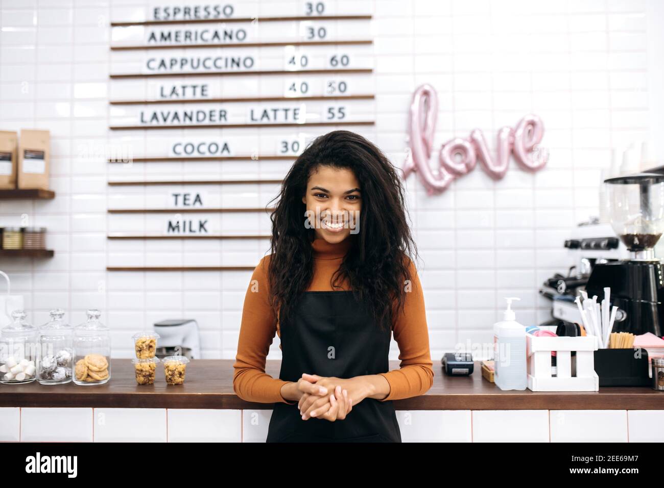 Kaffee-Business-Eigentümer Konzept. Attraktive junge multirassische schwarze weibliche Barista oder Kellnerin in schwarzer Schürze freundlich lächelt an der Kamera in der Nähe von Café-Theke Bar. Stockfoto