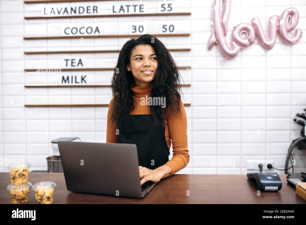 Hübsche junge afroamerikanische Kellnerin in schwarzer Schürze, Kleinunternehmerin, die am Laptop arbeitet, Online-Bestellungen verarbeitet, während sie hinter der Bar in ihrem Café steht und auf Besucher wartet Stockfoto