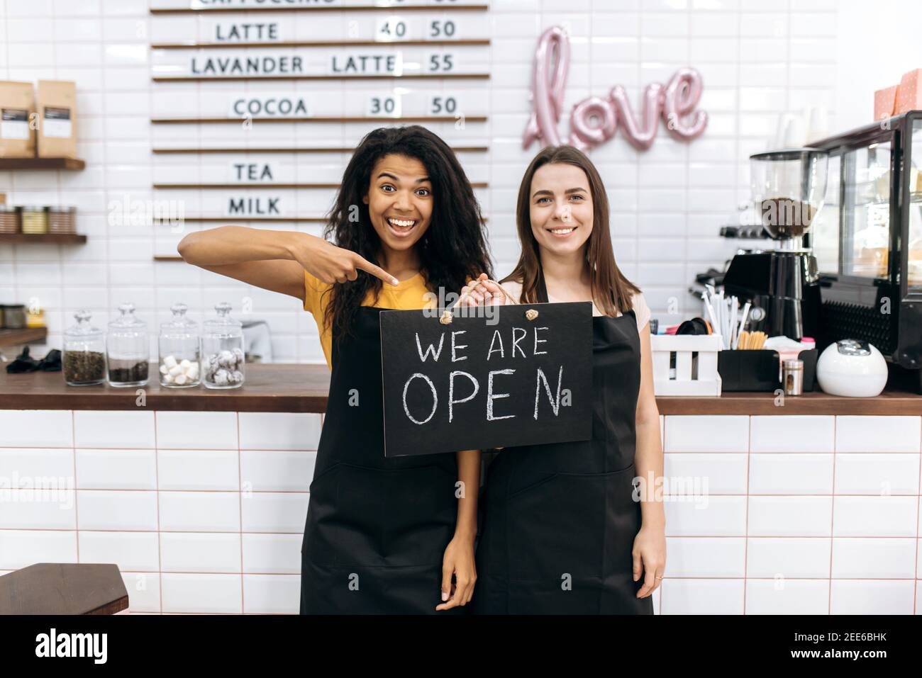 Zwei fröhliche weibliche Kellnerin ein Café oder Restaurant, afroamerikanisch und kaukasisch, steht mit einem Schild, DAS WIR IN der Nähe der Theke öffnen, Blick und lächelnd auf die Kamera Stockfoto