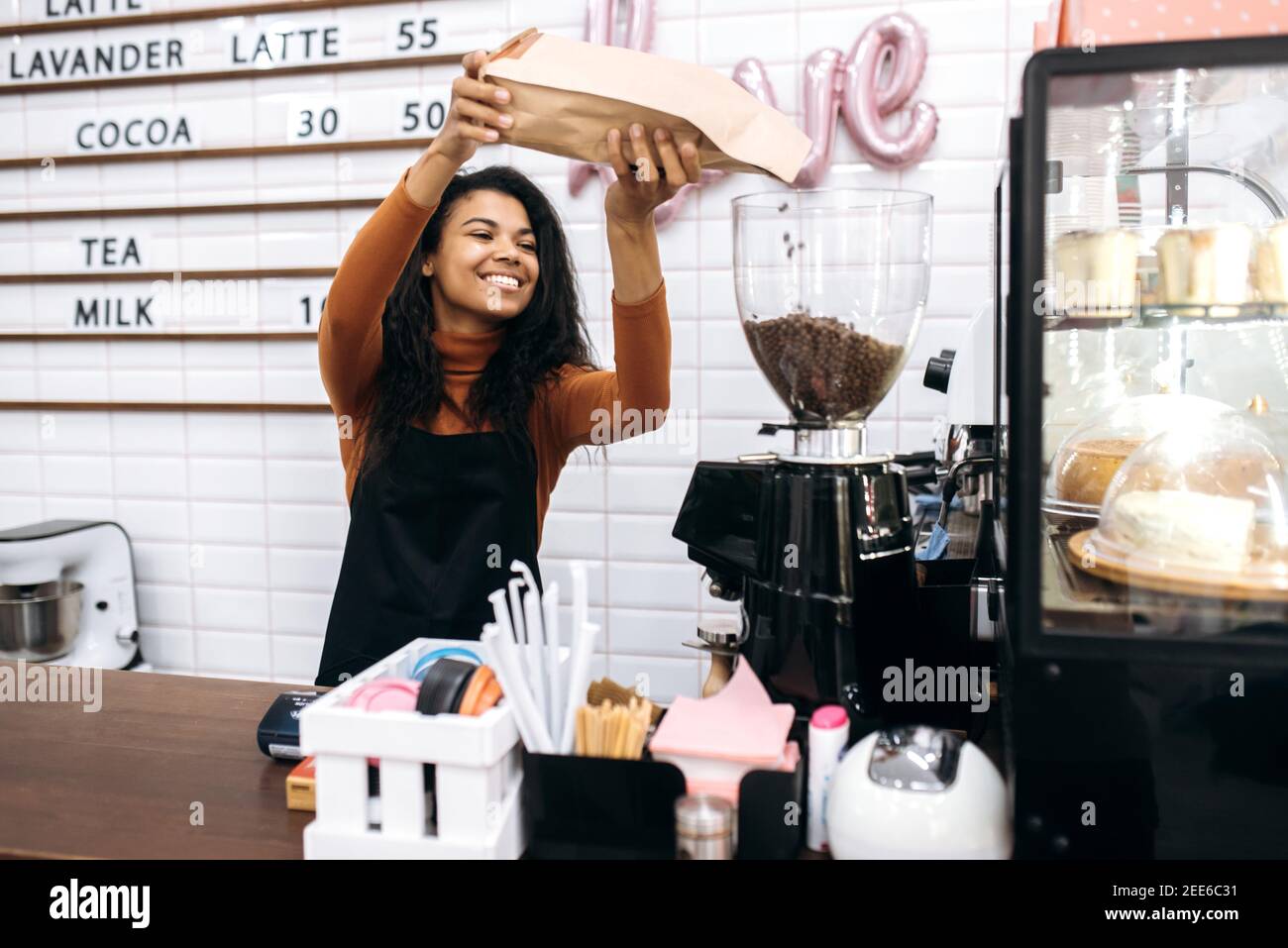 Lächelnde, multiethnische Barista in einer schwarzen Schürze gießt aromatische Kaffeebohnen in eine Kaffeemaschine. Konzept des Inhabers kleiner Unternehmen Stockfoto
