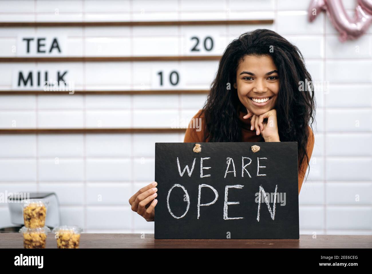 Porträt einer glücklichen multirassischen schwarzen Barista oder Kellnerin, Besitzer eines Coffee-Shop, steht mit einem Schild, DAS WIR HINTER der Theke ÖFFNEN, freundlich lächelnd und wartet auf Besucher Stockfoto