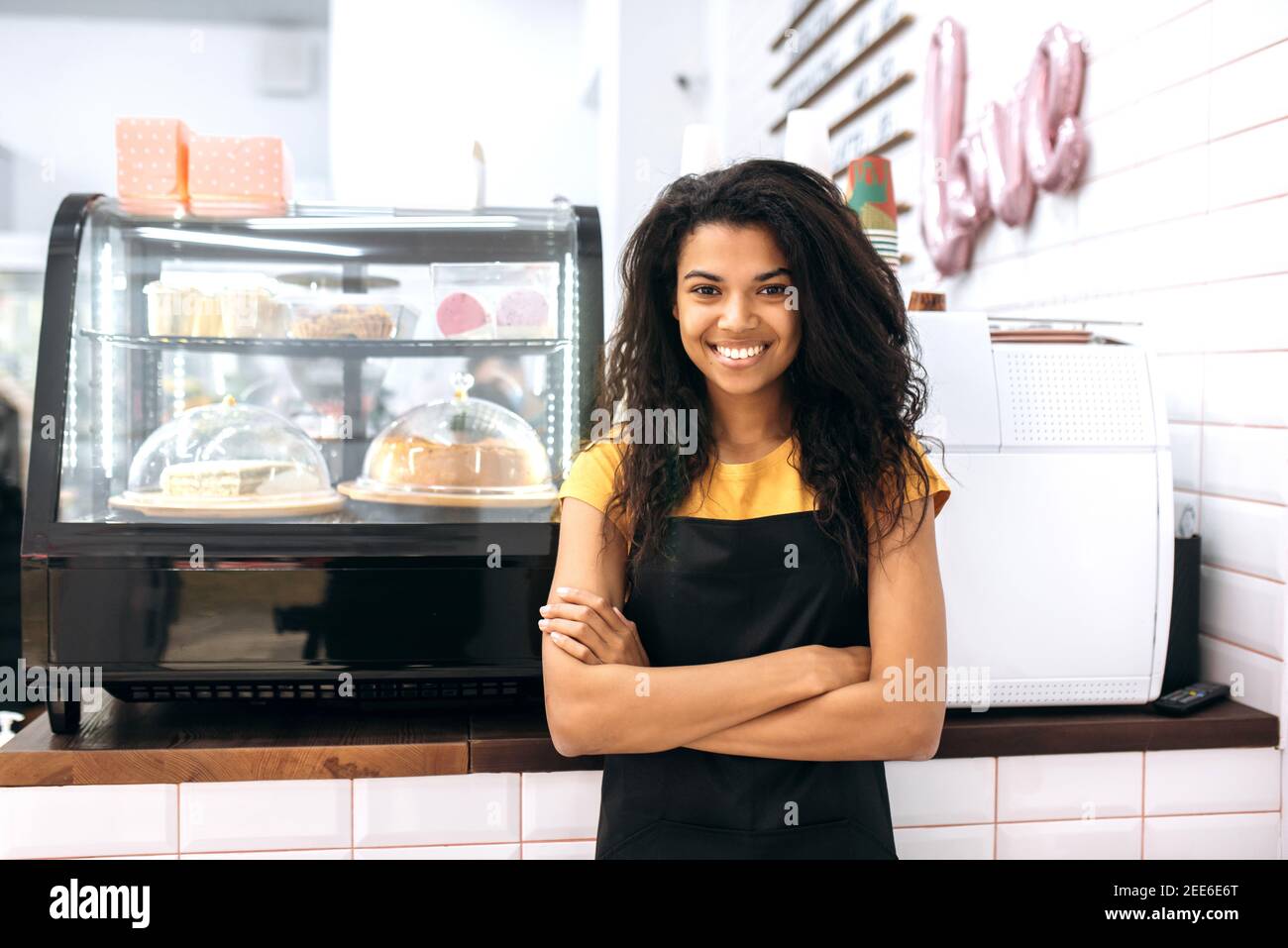Freundliches afroamerikanisches Mädchen, Kellnerin oder Geschäftsbesitzerin, steht in einem Café in der Nähe von Desserts, mit gekreuzten Armen, schaut und lächelt an der Kamera Stockfoto