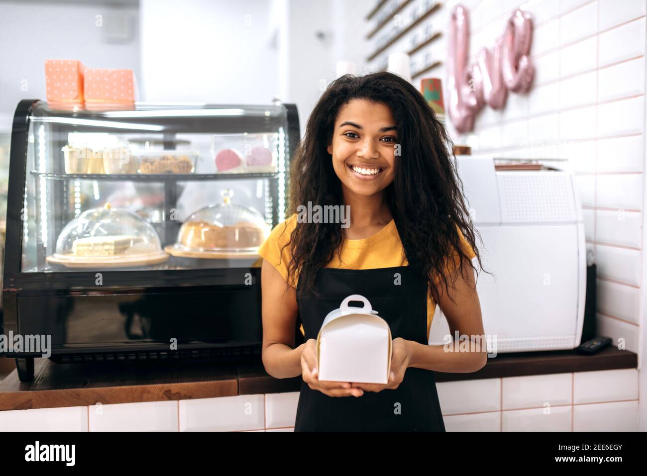 Zufriedene und freundliche multirassische schwarze Mädchen, Kellnerin oder Unternehmer, steht in einem Café, hält einen Karton mit Dessert innen, schaut und lächelt an der Kamera Stockfoto