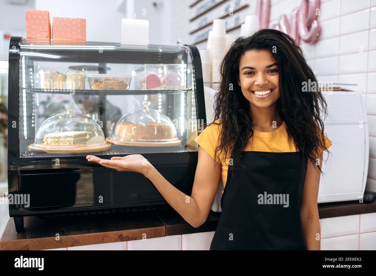 Freundliches afroamerikanisches Mädchen, Kellnerin oder Geschäftsinhaberin, steht in einem Café in der Nähe von Desserts, zeigt auf sie, schaut und lächelt auf die Kamera Stockfoto
