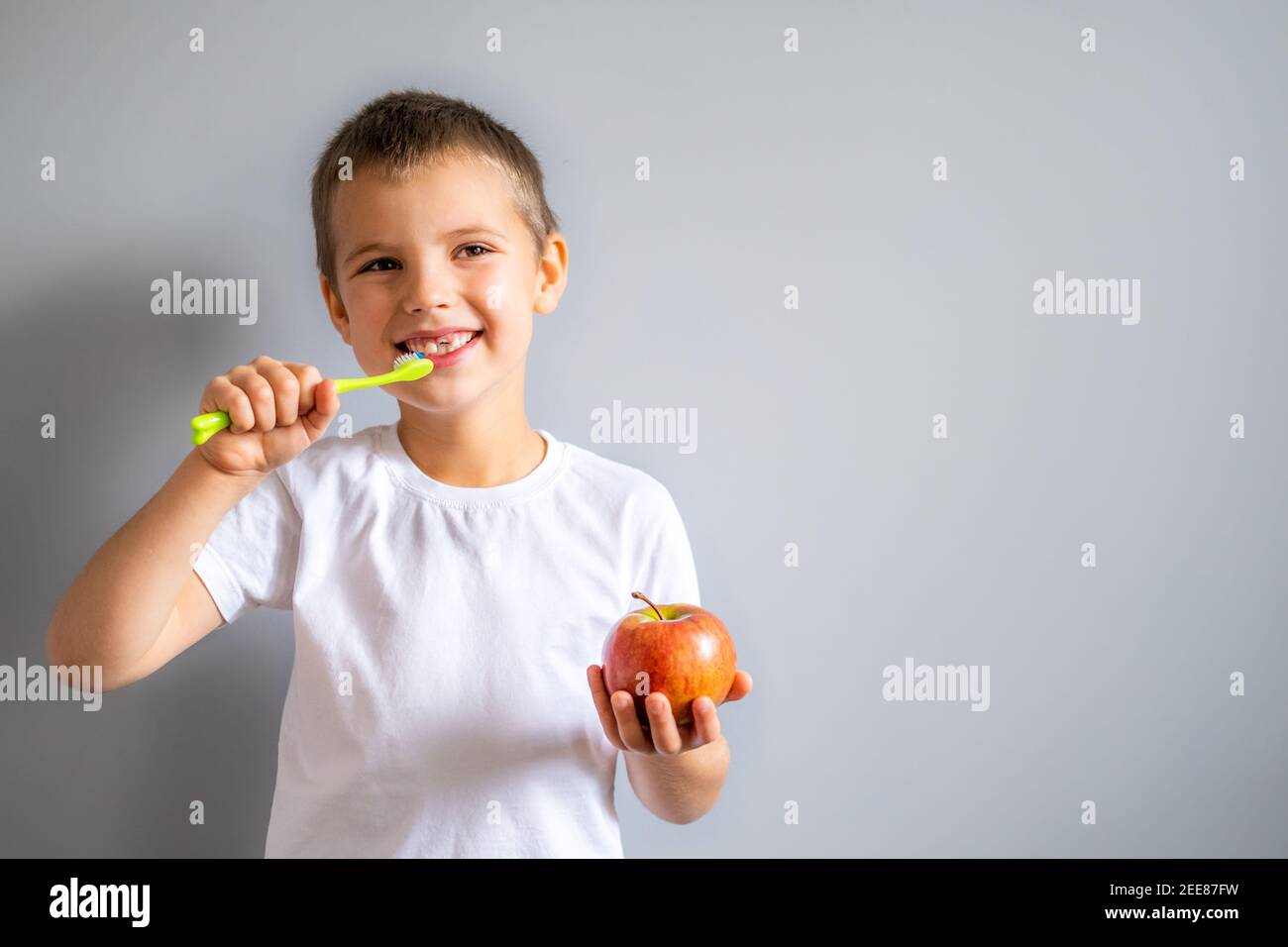 Junge ohne Milch Oberzahn in weißem T-Shirt lächelnd und hält einen Apfel und eine Zahnbürste in den Händen auf dem grauen Hintergrund. Stockfoto