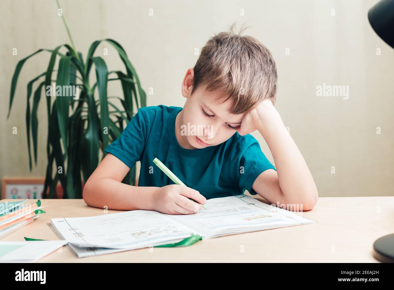 7 Jahre alter Junge, der am Schreibtisch sitzt und Hausaufgaben macht. Kind schreibt Notizen im Notizbuch, schwierige Hausaufgaben, Kind konzentriert sich auf Beispiele Stockfoto