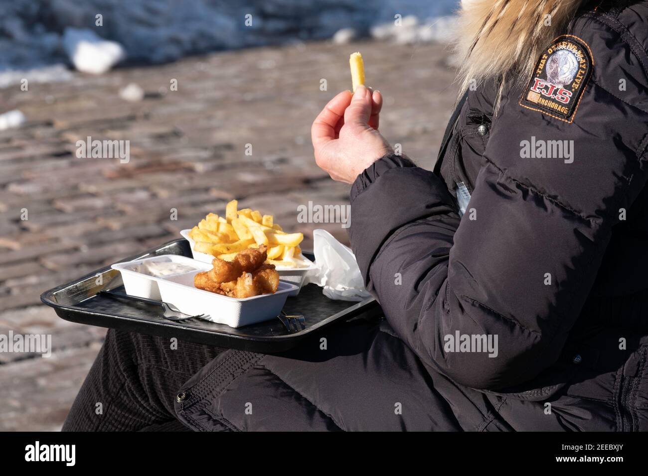 Frau in einem dicken Wintermantel isst Fisch und Chips, die draußen auf einer Bank auf einer Straße sitzen. Konzentrieren Sie sich auf die Schulter und die Fische Stockfoto