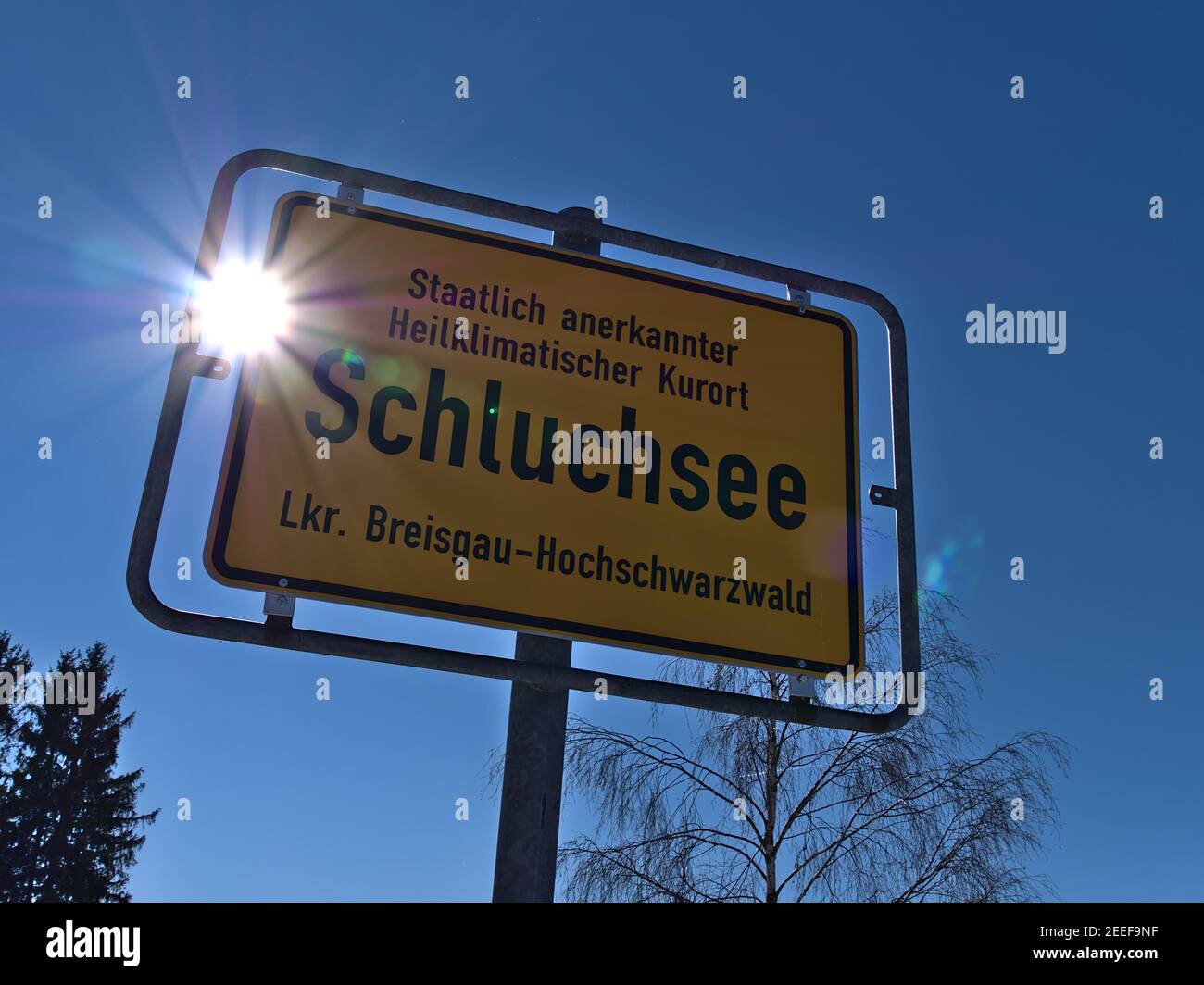 Nahaufnahme des gelbfarbenen Ortsnamenzeichens des Dorfes Schluchsee, einem staatlich anerkannten Kurort, im Gegenlicht mit Sonnenstrahlen und Fackeln. Stockfoto