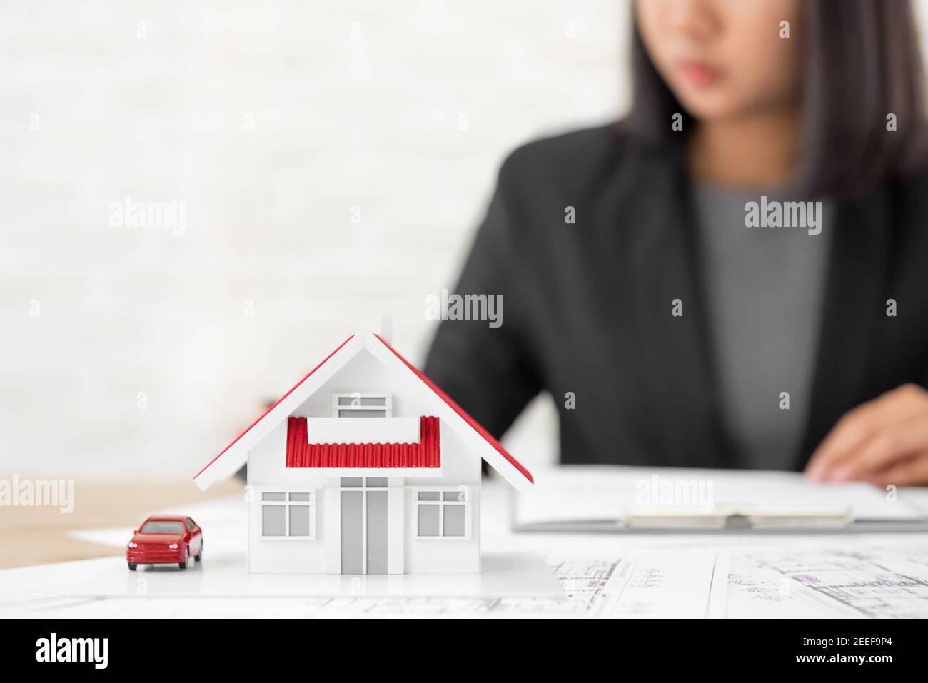 Hausmodell auf Blaupause Papier am Tisch mit verschwommen Geschäftsfrau im Hintergrund - Immobilien und Immobilienmakler Konzept Stockfoto