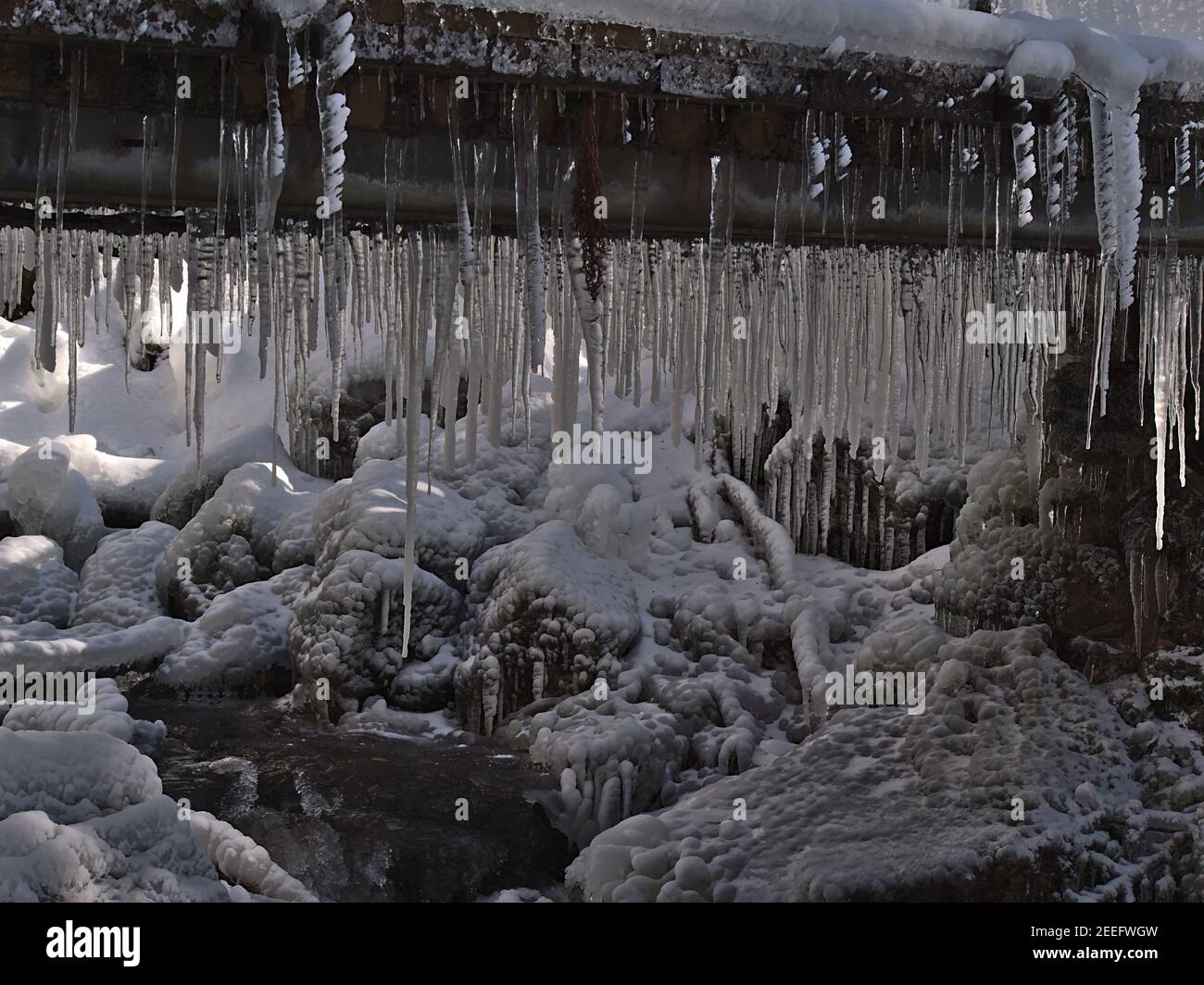 Viele durchsichtige Eiszapfen hängen von einer Fußgängerbrücke über einen Gebirgsbach mit gefrorenen Felsen im Flussbett bei Todtnauer Wasserfällen im Winter. Stockfoto