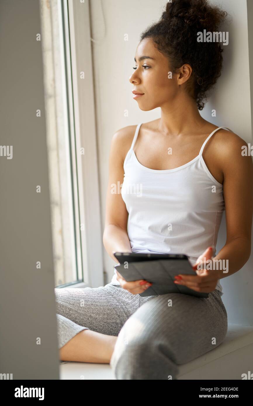 Nachdenkliche junge Frau mit Tablet-pc, die aus dem Fenster schaut und sich gemütlich fühlt, während sie alleine am Fenster zu Hause sitzt. Lifestyle, Wochenend-Konzept Stockfoto Nachdenkliche junge Frau mit Tablet-pc, die aus dem Fenster schaut und sich gemütlich fühlt, während sie alleine am Fenster zu Hause sitzt. Lifestyle, Wochenend-Konzept Stockfoto
