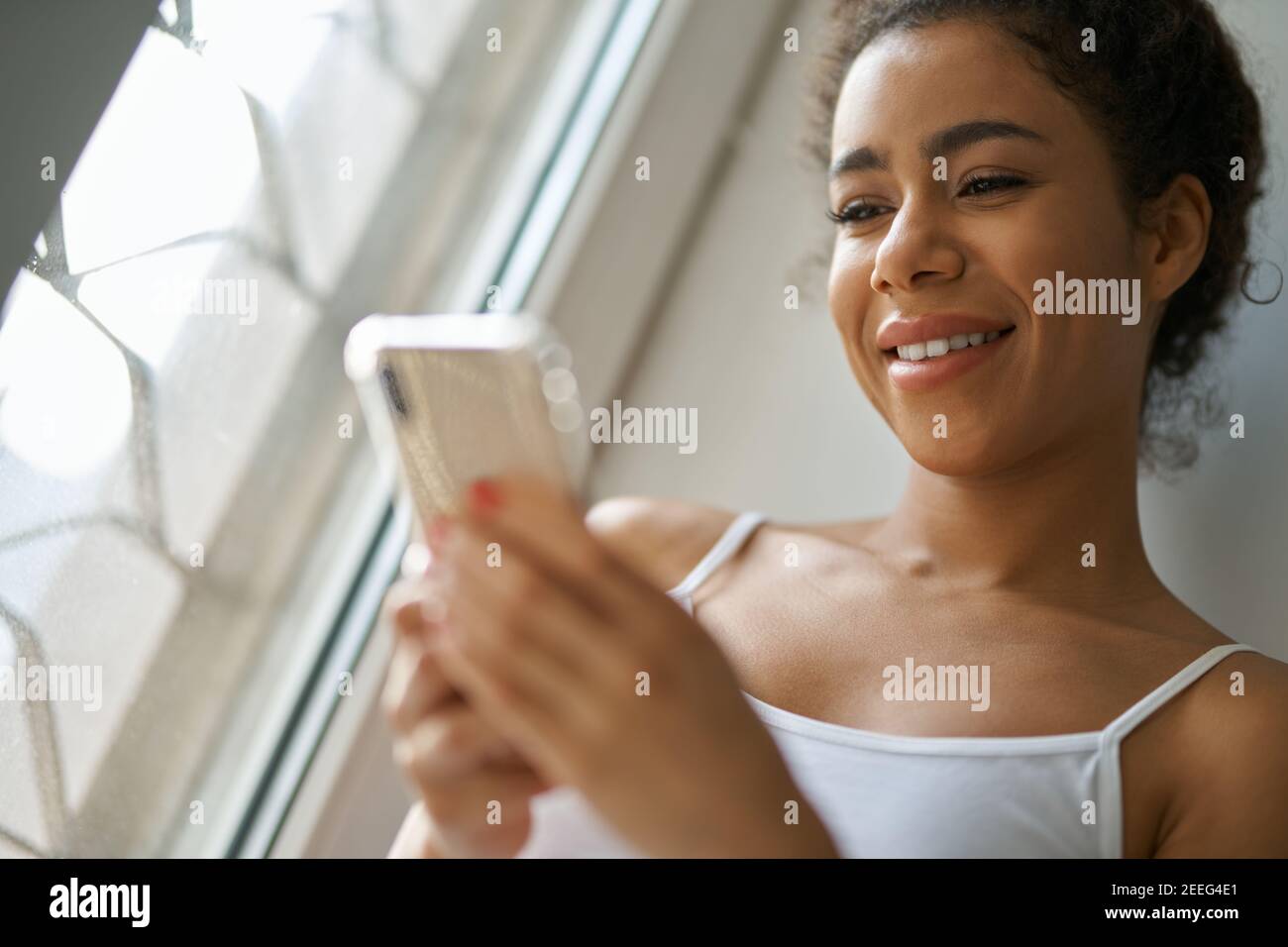 Porträt einer fröhlichen jungen gemischten Rasse Frau lächelnd, mit Smartphone, wohlig fühlen, während sie am Fenster zu Hause sitzen. Lifestyle, Freizeitkonzept Stockfoto Porträt einer fröhlichen jungen gemischten Rasse Frau lächelnd, mit Smartphone, wohlig fühlen, während sie am Fenster zu Hause sitzen. Lifestyle, Freizeitkonzept Stockfoto