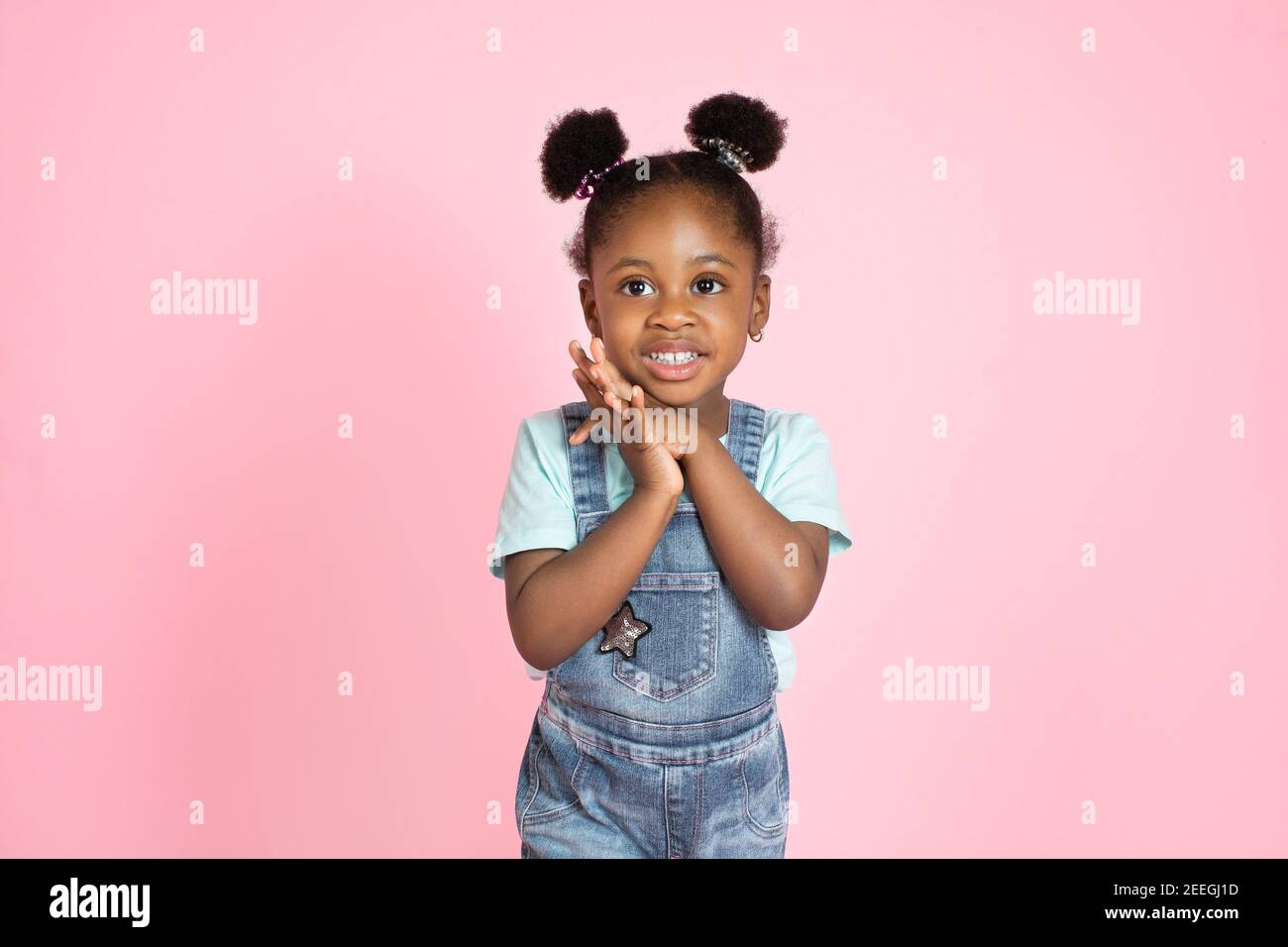 Glücklich lachende schwarze Haut kleines Mädchen in Freizeitkleidung, Jeans  Overalls, Blick auf die Kamera, die Hände zusammen halten. Kleines Kind mit  Afrohaar auf rosa Stockfotografie - Alamy