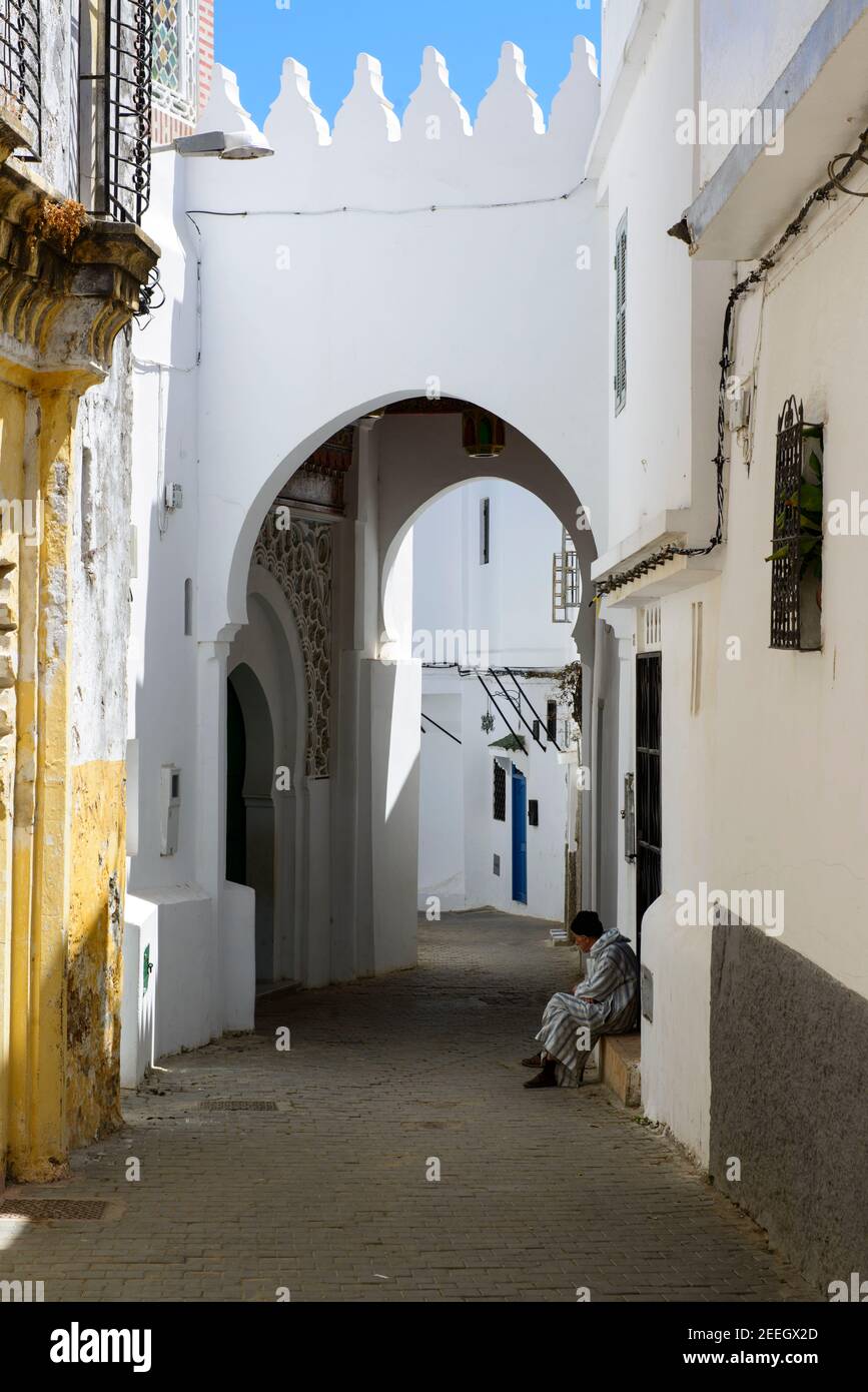 Ein Mann mit gestreifter Djellaba sitzt vor einer kleinen Moschee an der Schwelle eines Hauses in der Medina von Tanger, Marokko. Stockfoto