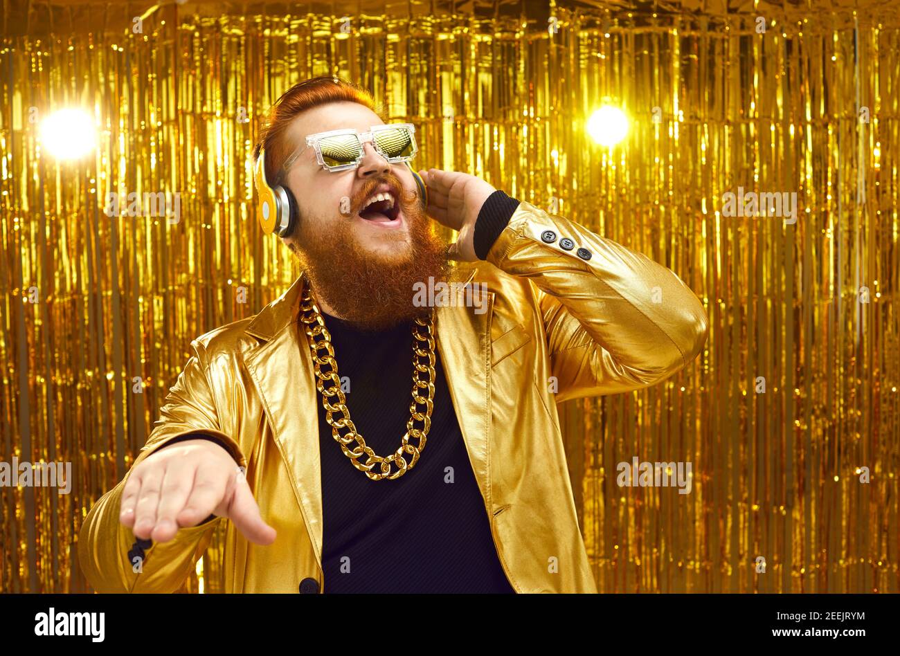 Fröhlicher DJ in goldglänzender Jacke, Kopfhörern und cooler Sonnenbrille, die laute Musik spielt Stockfoto