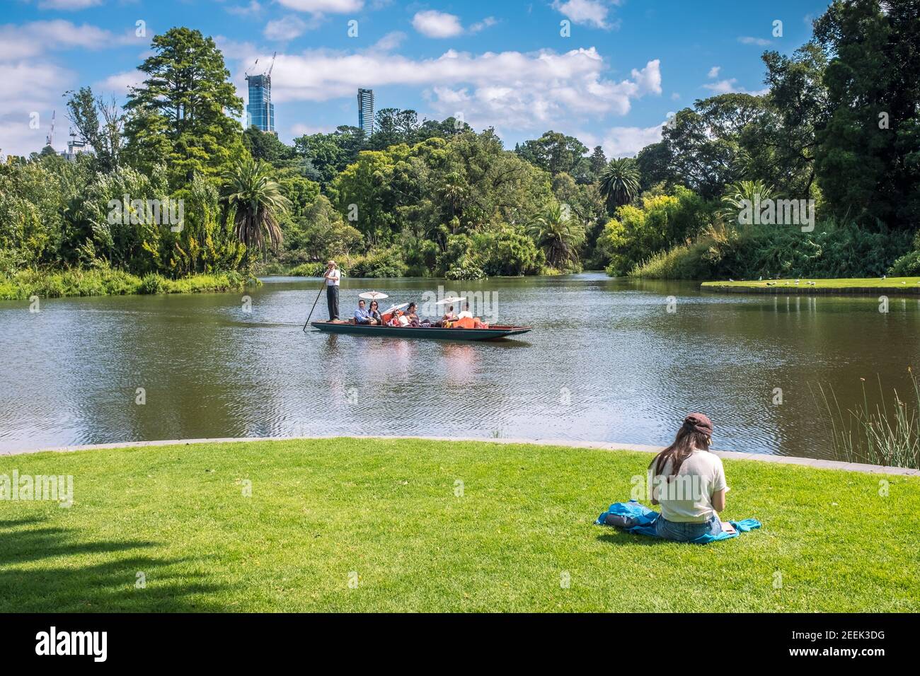 Eine Frau sitzt im Royal Botanic Gardens in Melbourne auf dem Gras, während eine Gruppe von Leuten in einem Punt im Hintergrund vorbeikommt Stockfoto