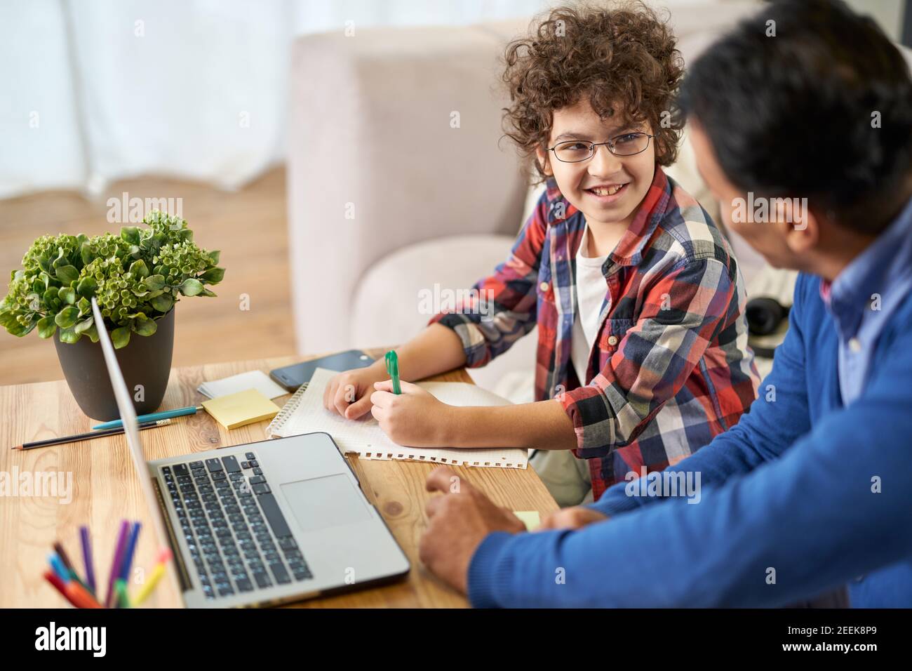 Porträt eines fröhlichen kleinen hispanischen Schuljungen, der zusammen mit seinem Vater Hausaufgaben macht und dabei Laptop benutzt, während er zu Hause am Schreibtisch sitzt. Online studieren, Familie, Vaterschaft Konzept Stockfoto