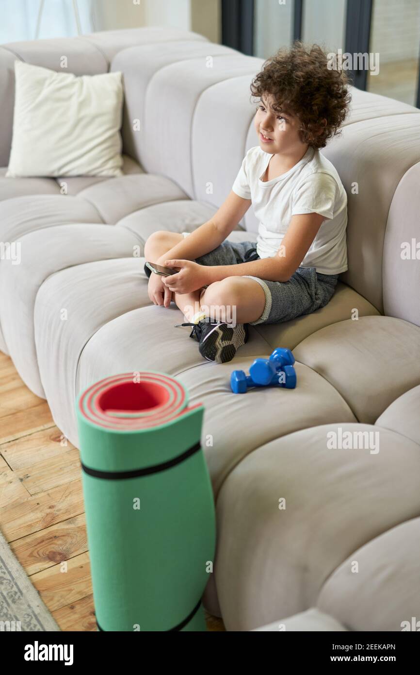 Netter lateinischer Junge, der auf einem Sofa sitzt und Smartphone benutzt, anstatt Sport zu treiben, während er Zeit zu Hause verbringt. Freizeit, Kinder, Technik Konzept Stockfoto