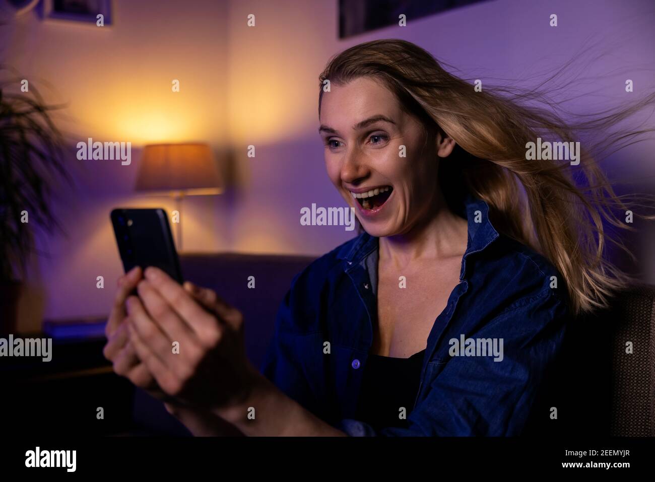 Schockiert überrascht junge Frau auf der Couch sitzen und mit Handy. Blasen Haare Stockfoto