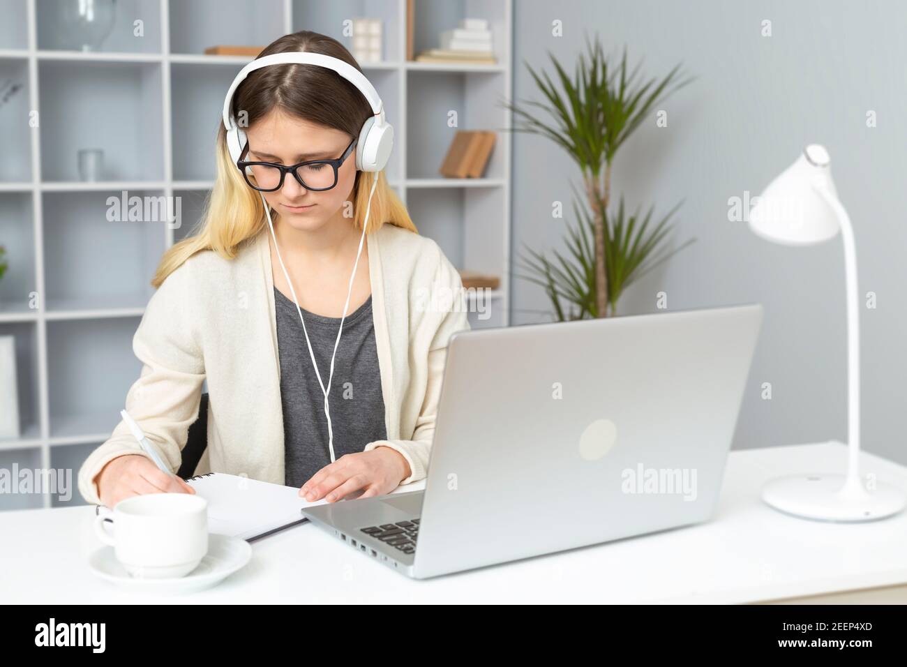 Mädchen Schülerin in Kopfhörer hört sich die Lehrer-Unterricht online mit einem Laptop. Eine junge Frau mit Brille, die zu Hause sitzt, schreibt einen Vortrag in einer Notiz Stockfoto