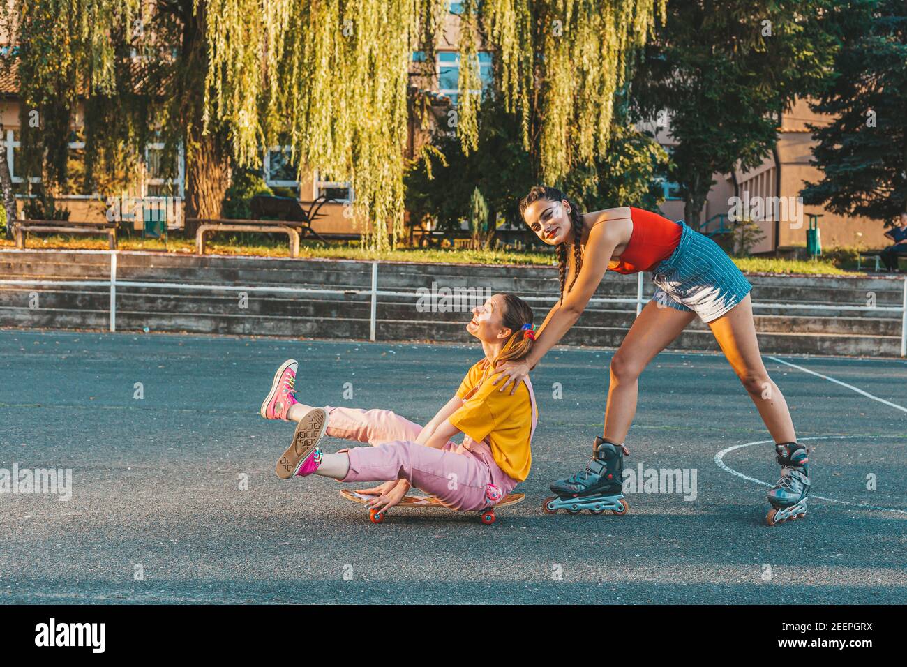Zwei Teenager-Mädchen, Freunde, die Spaß mit Skate-Board. Ein Mädchen schiebt andere von hinten Stockfoto