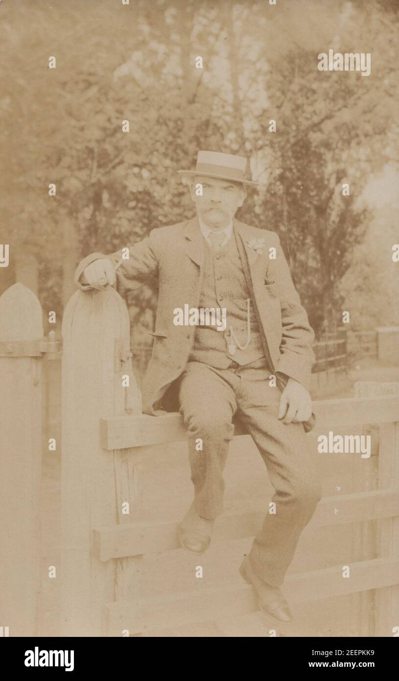 Vintage Fotokarte aus dem frühen 20. Jahrhundert zeigt einen suited man, der einen Bootshut trägt, während er auf einem hölzernen Tor saß. Stockfoto