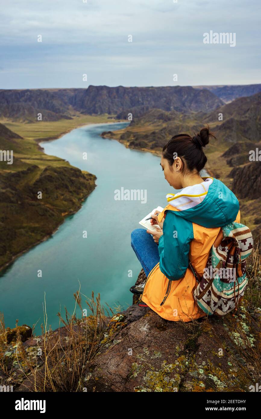 Eine junge Künstlerin zeichnet eine Frühlingslandschaft eines herrlichen Canyon in der Natur. Das Mädchen sitzt auf einem Stein am Fluss. Lackierung im Freien. Der Ili River Stockfoto